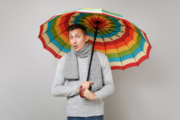 Shocked young man in gray sweater, scarf holding colorful umbrella isolated on grey background in...