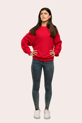 A full-length shot of a Teenager girl with red sweater with confuse face expression while bites lip over isolated background