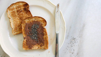 Two slices of toast bread with chocolate spread and chia seeds sprinkled on top, placed on a plate with bread knife, on marble countertop.