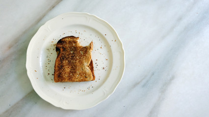 Toast bread bitten off at the top corner, on a white plate with crumbs around, on top of marble counter.