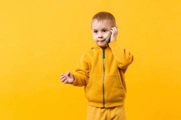 Little cool kid boy 3-4 years old in yellow clothes talking on mobile phone isolated on orange wall background, studio portrait. Problem of children and gadgets. Lifestyle concept. Mock up copy space.
