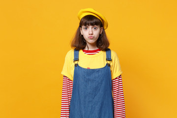 Portrait of confused cute girl teenager in french beret and denim sundress blowing lips isolated on yellow wall background in studio. People sincere emotions, lifestyle concept. Mock up copy space.