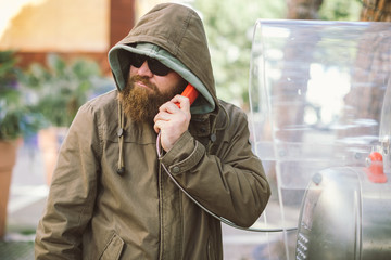 Portrait of young bearded man using public phone wearing hoodie (jacket) and black sunglasses -...