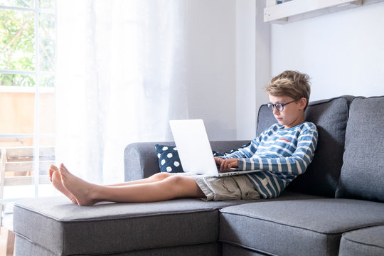 Beautiful blond child sitting on the couch at home using computer. Young boy relaxing with laptop free time online games. Teen working and chat with internet friends. Student doing homework on the web