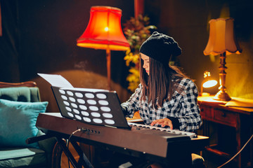 Beautiful young Caucasian woman with hat on head sitting and playing clavier in home studio.