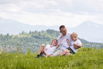 Happy father with his two young sons sitting on the grass on a background of green forest, mountains and sky with clouds. Friendship concept.