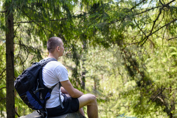 Naklejka na ściany i meble Young man with a backpack sits on a rock in the coniferous forest. Side view