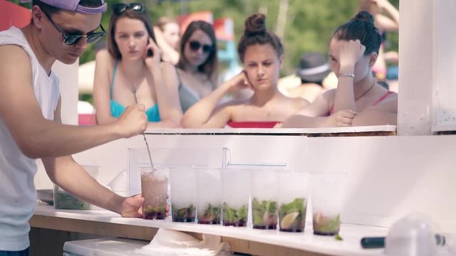 4k shot: Young girls stand near a bar on the beach, waiting for the bartender to prepare summer refreshing cocktails for them, with ice, berries and fruits.