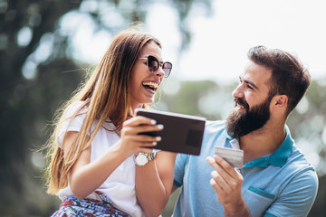 Cheerful young couple sitting on a park bench and uses a digital tablet for online shopping