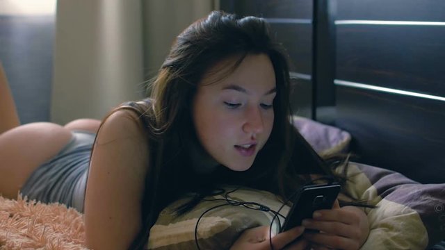 Young brunette woman in bed uses the phone. A girl enjoys listening to music.