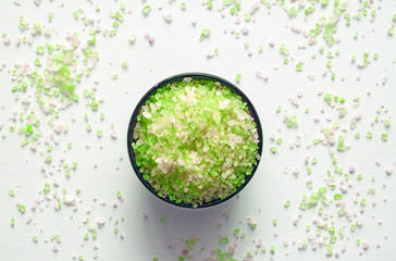 The fragrant and revitalizing sea salt for a bath of green and white in a brown bowl stands on a white table in the center, with salt crystals scattered around it.   Close up on a soft natural