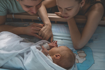 Woman and man look at newborn. Boy sleeps in his crib. Mom, dad and baby. Portrait of young family....