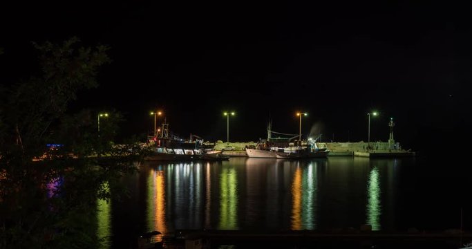 Timelapse night shot of quay with moored boats and people walking the pier. Colored lantern lights reflecting in dark water, Greece