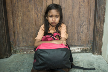 7 or 8 years child in school uniform sitting outdoors on the floor crying sad and depressed holding her backpack suffering bullying and abuse problem feeling helpless