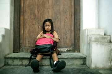 7 or 8 years child in school uniform sitting outdoors sad and depressed with her backpack on the stairs suffering bullying and abuse problem feeling alone and helpless