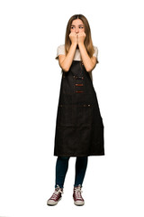Fototapeta premium Full body Young woman with apron is a little bit nervous and scared putting hands to mouth on isolated background