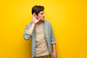 Teenager man over yellow wall listening to something by putting hand on the ear