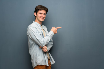 Teenager man with jean jacket over grey wall pointing finger to the side