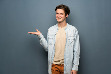 Teenager man with jean jacket over grey wall holding copyspace imaginary on the palm to insert an ad