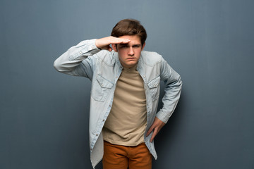 Teenager man with jean jacket over grey wall looking far away with hand to look something