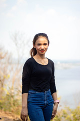 Women white skin lovely brown hair a small woman red lips wearing a black shirt wearing a blue gene woman stand  poses photography portrait which has both trees and water in the back in the park.