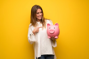 Blonde woman over yellow wall holding a piggybank