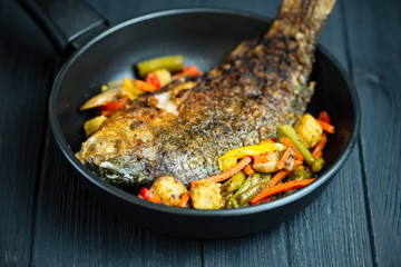 fried fish carp in a pan with vegetables on a wooden background