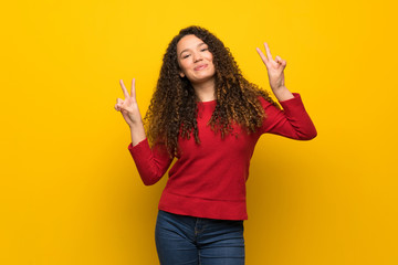 Teenager girl with red sweater over yellow wall smiling and showing victory sign with both hands