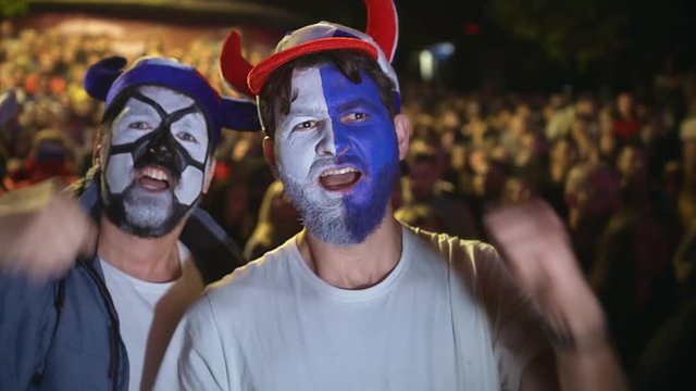 Football Fan Shouts, Rejoices Goal Favorite 4K Team. Man Paint Face Jump In Delight Victory Match. Closeup Guy Screaming Furiously, Jumping With Friend Against Backdrop Crowd Winning Football Game