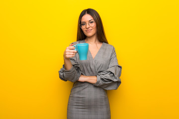 Young woman with glasses over yellow wall holding a hot cup of coffee