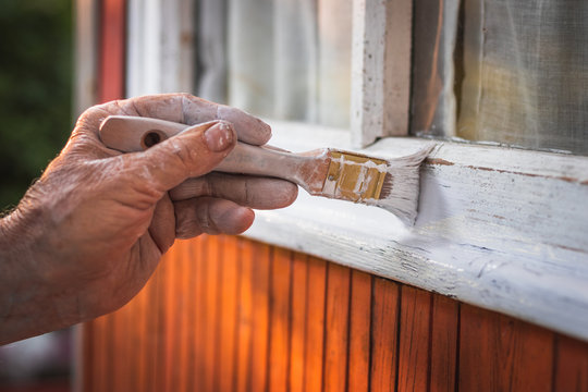 Old man´s hand painting wooden window frame using paintbrush. Repairing exterior of wooden hut