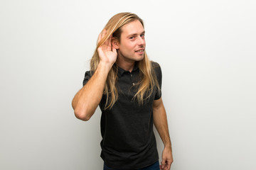 Blond man with long hair over white wall listening to something by putting hand on the ear