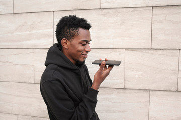portrait smiling african man holding in hands smartphone using voice command recorder, assistant or calling, looking at phone walking on city street on gray wall background