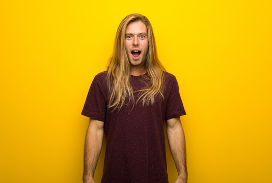 Blond man with long hair over yellow wall with surprise and shocked facial expression
