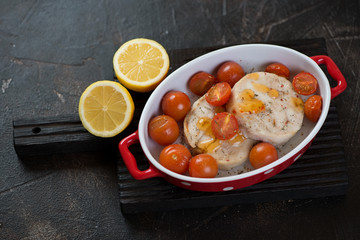 Baked fish medallions with cherry tomatoes and lemon over dark brown stone background, studio shot
