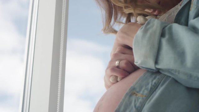 A beautiful pregnant girl in a blue jacket stands at the window and hugs a toy bear. Beautiful makeup and hairstyle. Pregnant belly close-up. Expecting a baby. Slow motion. 4K