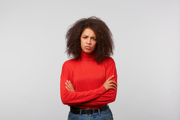Serious suspicious latino woman with curly afro hair standing with crossed hands and frowning, expressing distrustfulness and disappointment, over white background