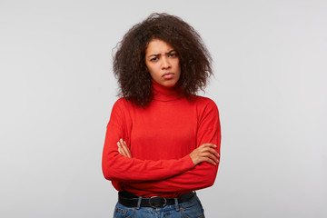 Indoor photo of serious suspicious latino woman with curly afro hair standing with crossed hands and frowning, expressing distrustfulness and disappointment, over white background