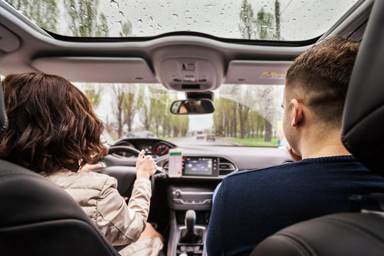 Beautiful Couple Sitting On The Front Passenger In Vehicle. Woman Is Driving A Car. View From A Back Of Young Pair. Travel And Adventure Concept