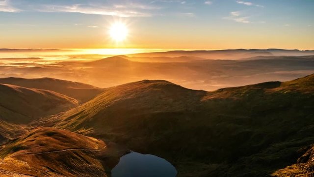 A time-lapse of a stunning orange yellow sunrise in Spring Summer from Sharp Edge on Blencathra, Lake District, Cumbria with reflection in lake tarn and cloud inversion over mountains in the distance
