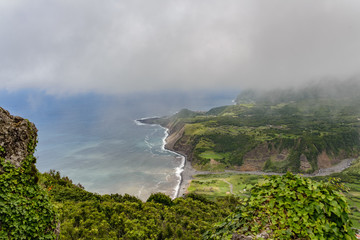 A beach on the island of Flores, in the Azores. It is seen from above, with plants in the foreground, low cloud, and the blue ocean.