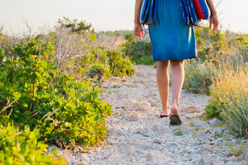Close up shot of young woman legs wearing flip flops and summer clothes carrying beach sleeping pad while walking on a trail. Summer holidays and travel concept.