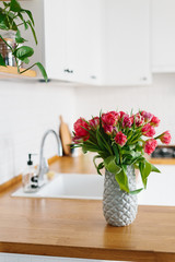 Tulips bouquet in vase standing on wooden countertop in the kitchen. Modern white u-shaped kitchen in scandinavian style.
