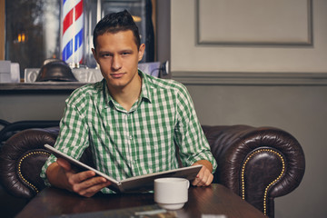 Handsome young man in a barbershop is waiting for the master in the waiting room while drinking coffee