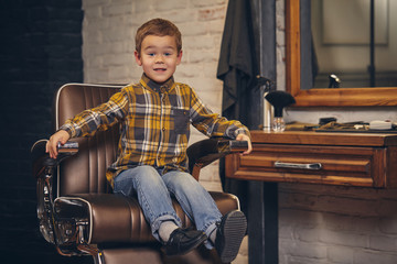 Portrait of a stylish little boy dressed in shirt and jeans in the barbershop, sitting in a chair...