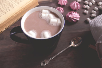 Obraz na płótnie Canvas Сocoa coffe with marshmallows in a dark blue cup on a wooden table next to a book, a spoon and meringue atmospheric view