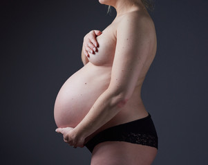 A beautiful pregnant woman body, with the woman gently holding and supporting the pregnant stomach. Gentle feminine motherhood. Photographed on a black studio background.