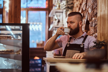 Handsome barista in apron drinking coffee during lunch break sitting at a table in the coffee shop
