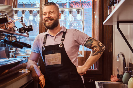 Confident barista with stylish beard and hairstyle wearing apron smiling and looking sideways while leaning on a counter in the cafe or restaurant