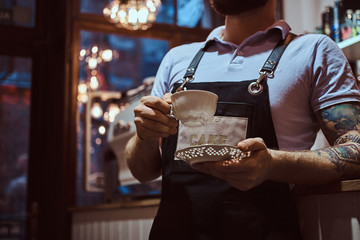 Barista wearing apron holding a cup and saucer, drinking coffee during lunch break leaning on a counter in the coffee shop. Cropped image